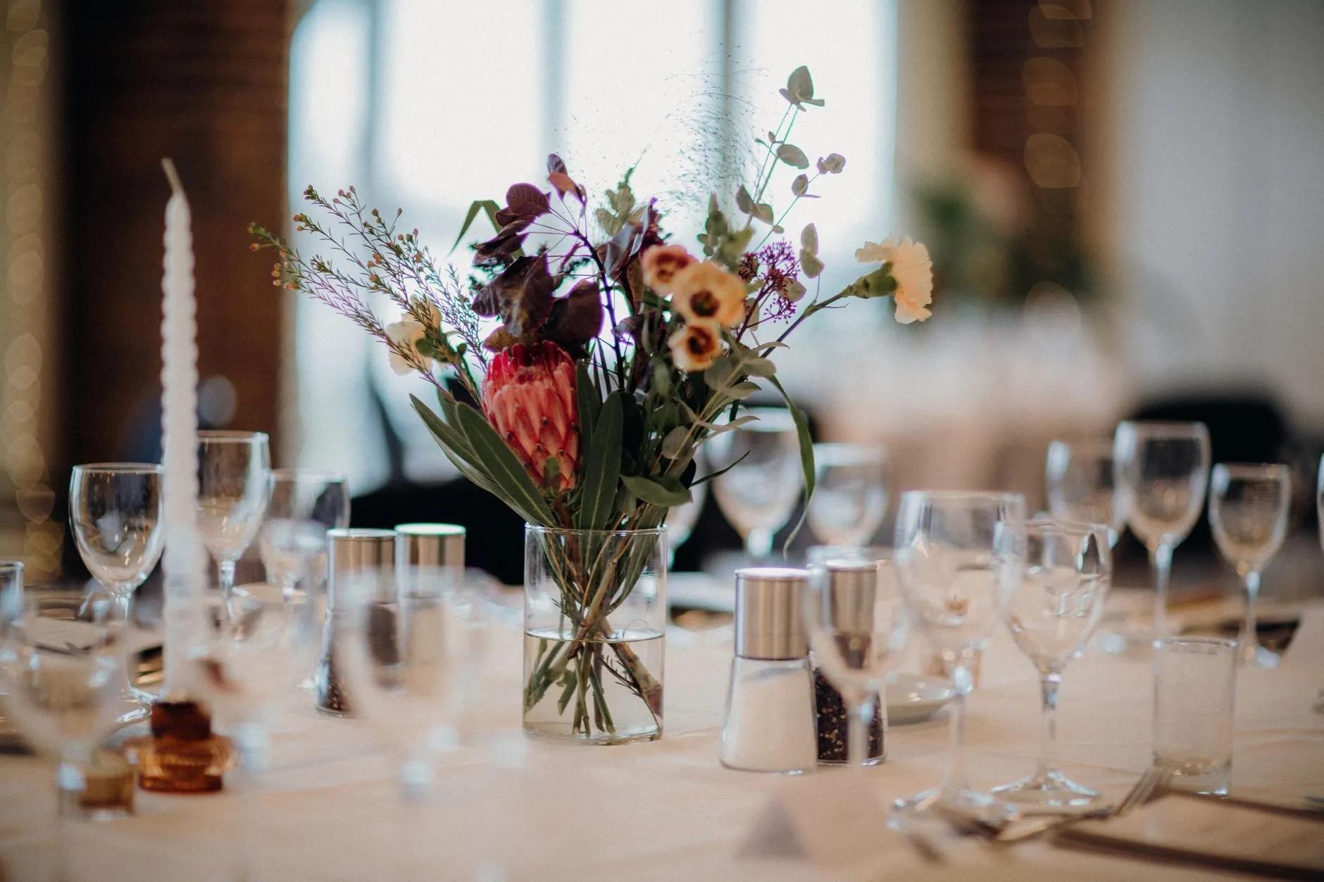 A reception table set with glasses, a tall taper candle and a loose arrangement in a glass vase